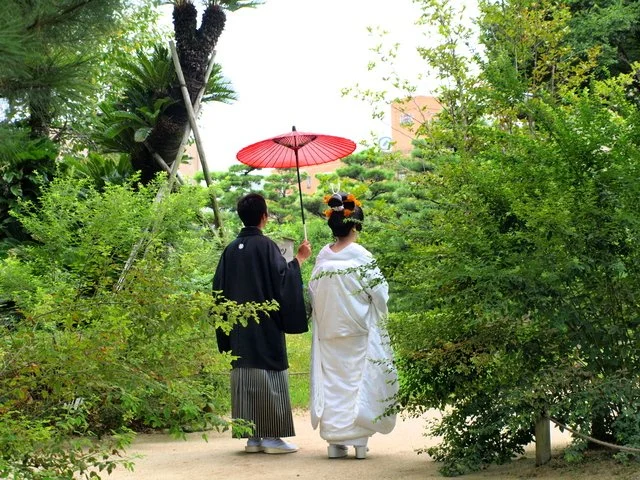 緑豊かな庭園で、赤い和傘をさして歩く白無垢と紋付袴の二人